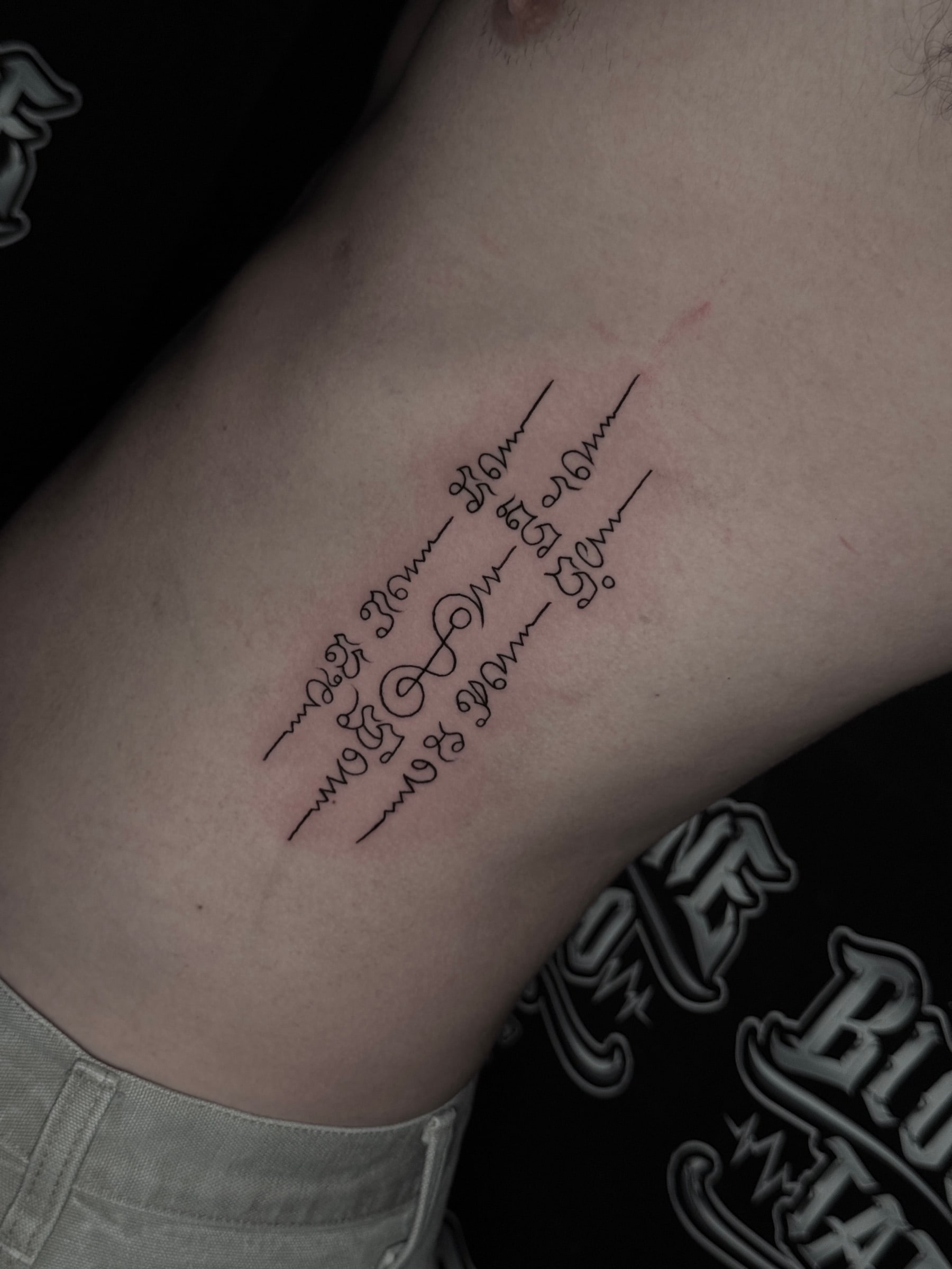 Three-line Sak Yant with diagonal Khom script columns and unalome symbols on the side ribcage at Bloodline Tattoo Phuket.