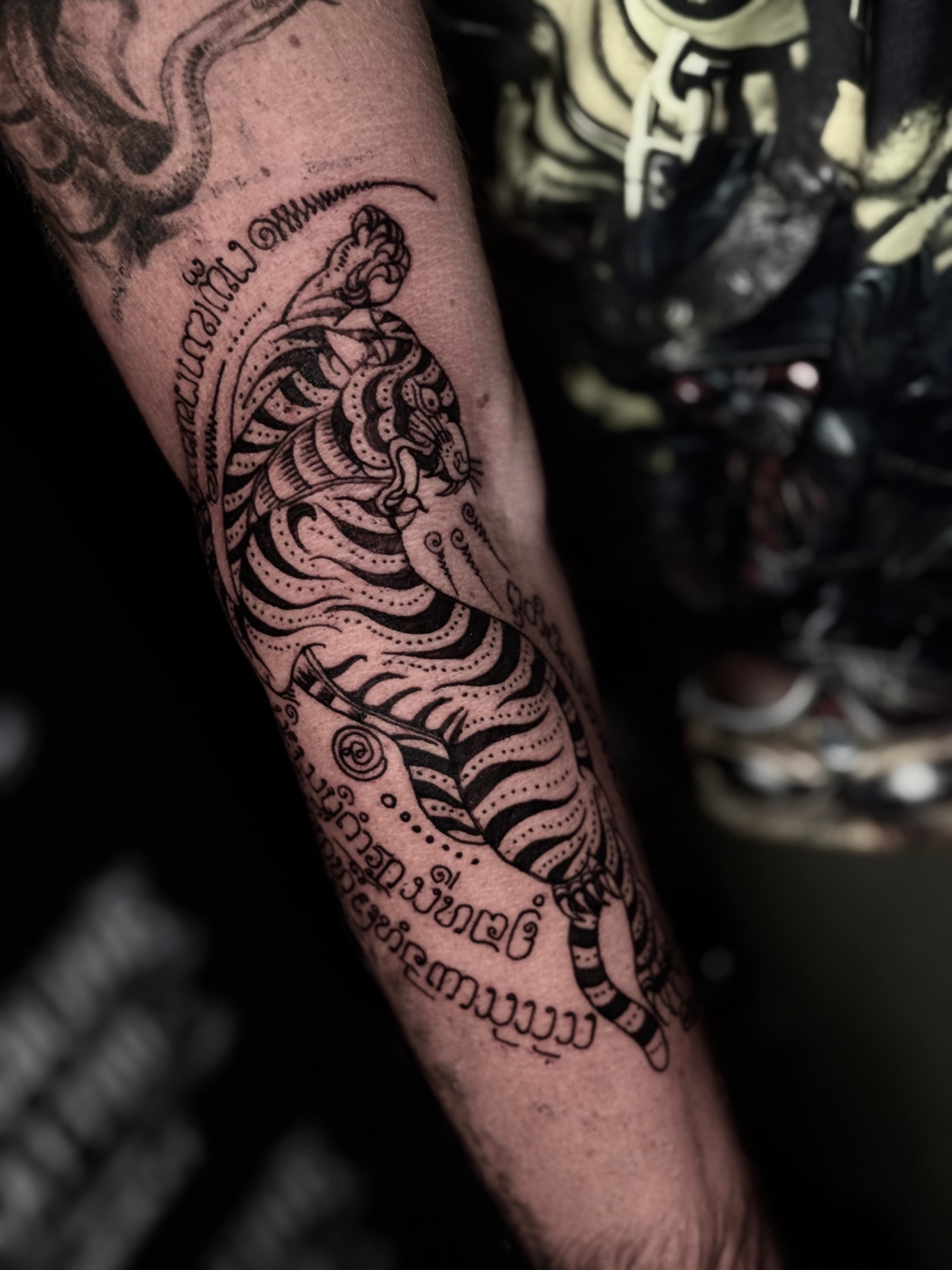 Suea (Tiger) Sak Yant of a leaping tiger with Khom script arcs and unalomes on the inner forearm at Bloodline Tattoo Phuket.
