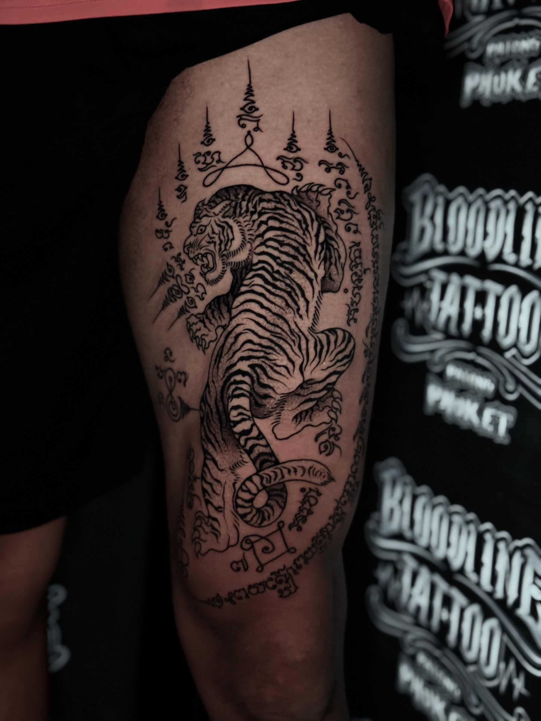 Suea (Tiger) Sak Yant of a snarling tiger with Khom script border and multiple unalomes on the outer thigh at Bloodline Tattoo Phuket.