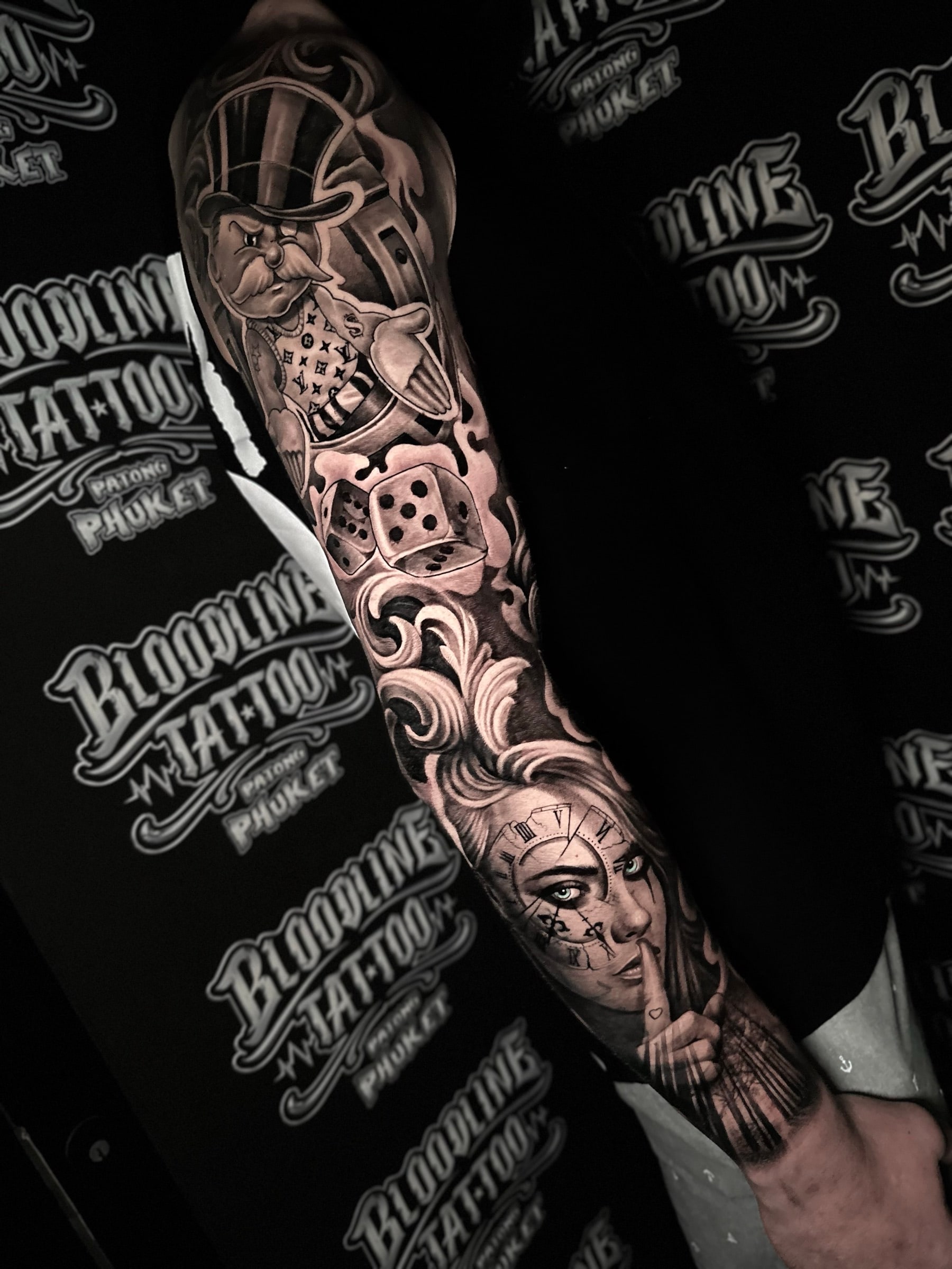 Chicano black and grey sleeve with a Monopoly man in an LV-pattern vest, tumbling dice, and a woman portrait with a clock eye and shush gesture at Bloodline Tattoo Phuket.