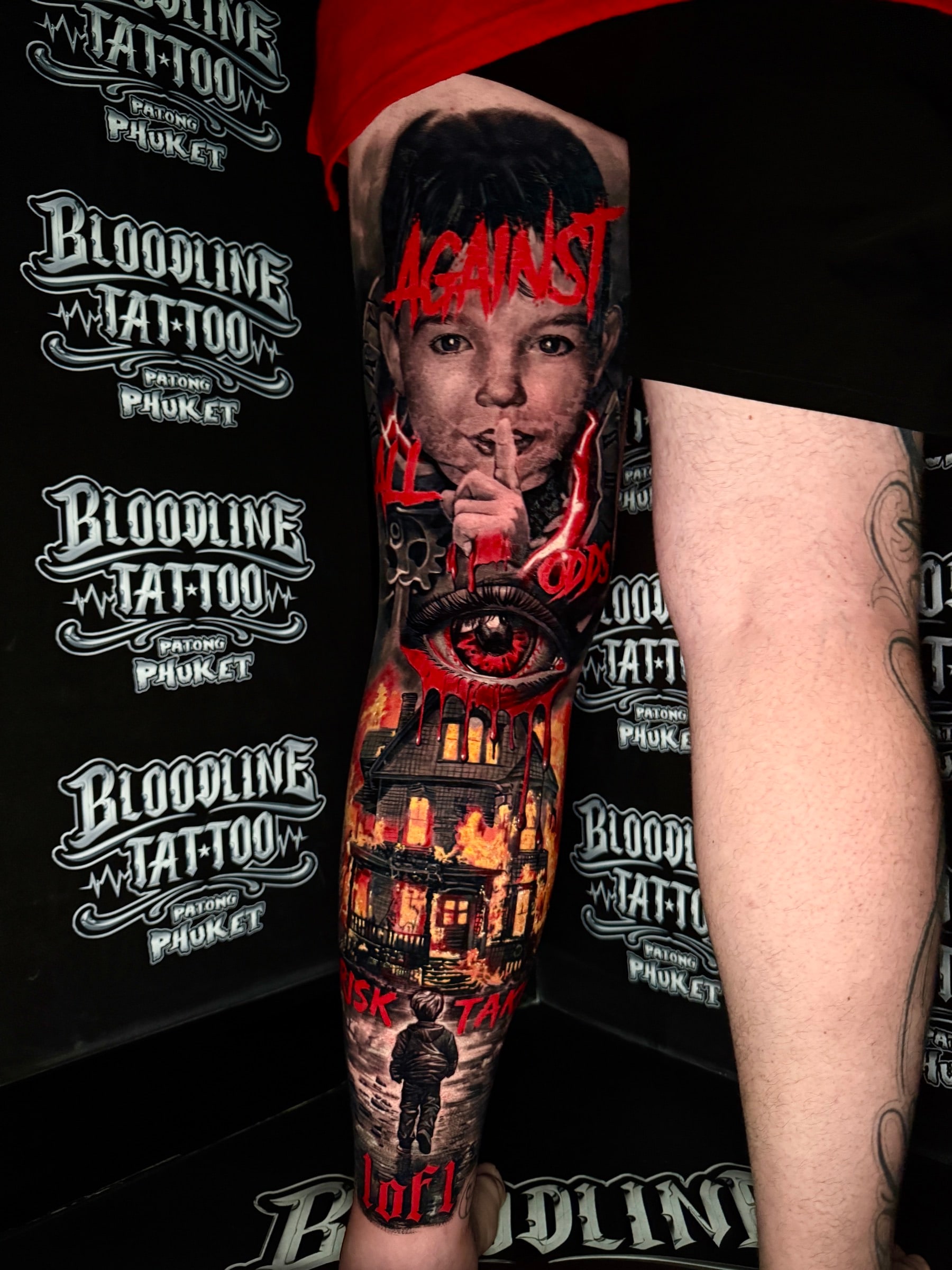 Trash-polka colour leg tattoo with a boy portrait, AGAINST ALL ODDS script, a red crying eye, a burning house, and RISK TAKER lettering at Bloodline Tattoo Phuket.
