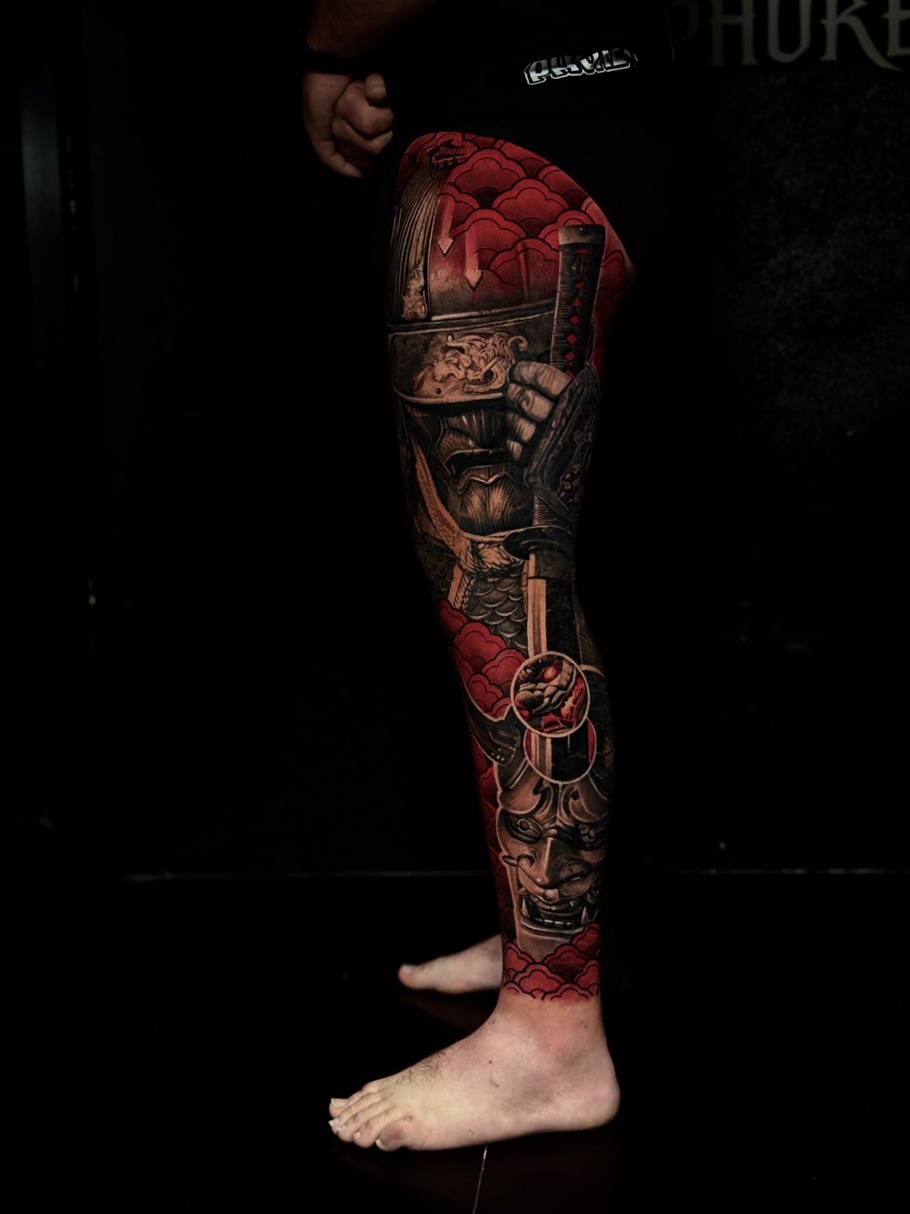 Japanese-inspired colour leg tattoo of an armored samurai with a katana and a Hannya mask amid red cloud accents at Bloodline Tattoo Phuket.