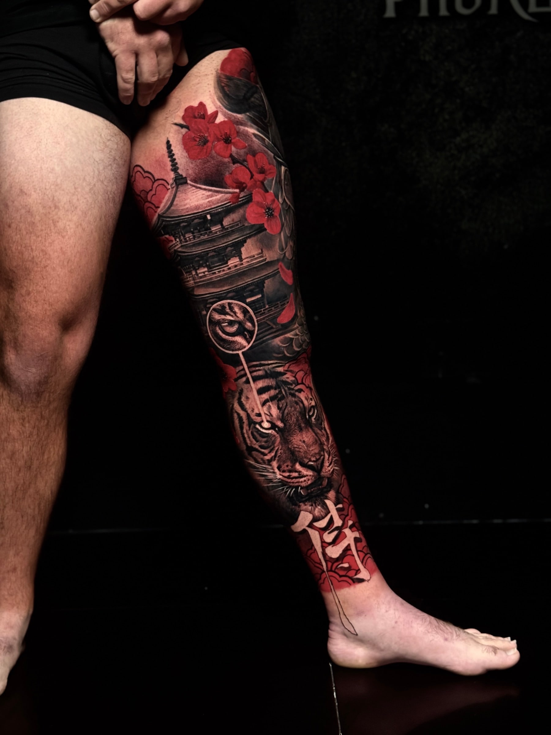 Japanese colour leg tattoo with a pagoda, red cherry blossoms, a tiger eye medallion, a realistic tiger portrait, and kanji at Bloodline Tattoo Phuket.