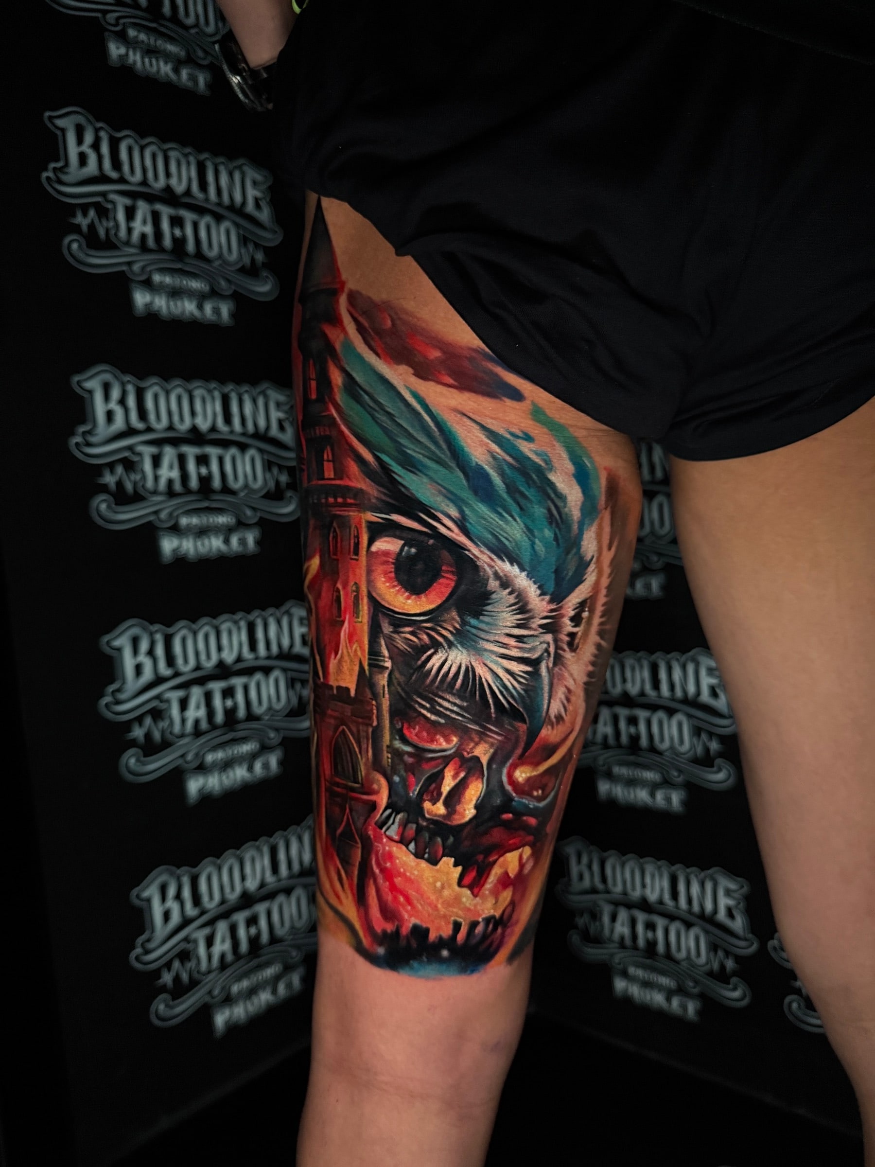 Surrealist colour thigh tattoo of a large orange owl eye, a stylized skull, and a Gothic tower engulfed in flames at Bloodline Tattoo Phuket.