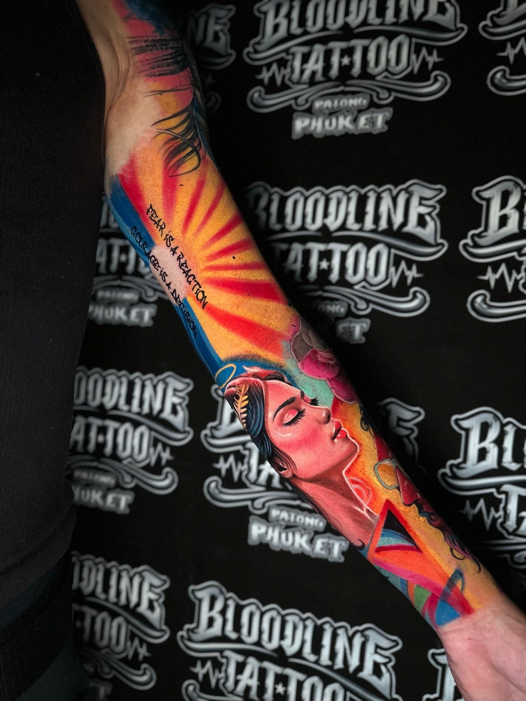 Colour forearm tattoo of a laurel-crowned woman profile with FEAR IS A REACTION and COURAGE IS A DECISION script beside a sunburst at Bloodline Tattoo Phuket.