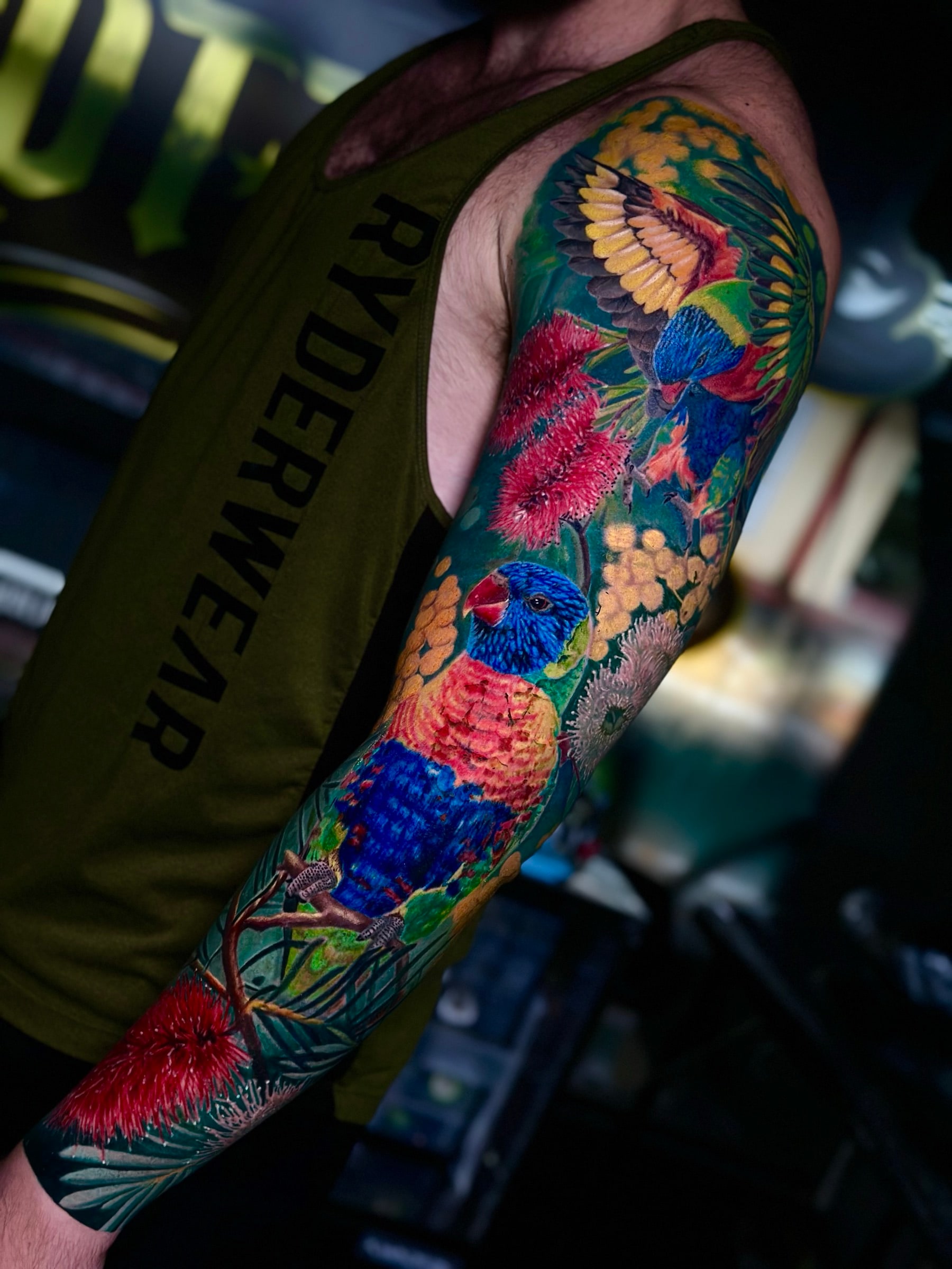 Australian colour sleeve with rainbow lorikeets, red bottlebrush flowers, and golden wattle on dark green foliage at Bloodline Tattoo Phuket.