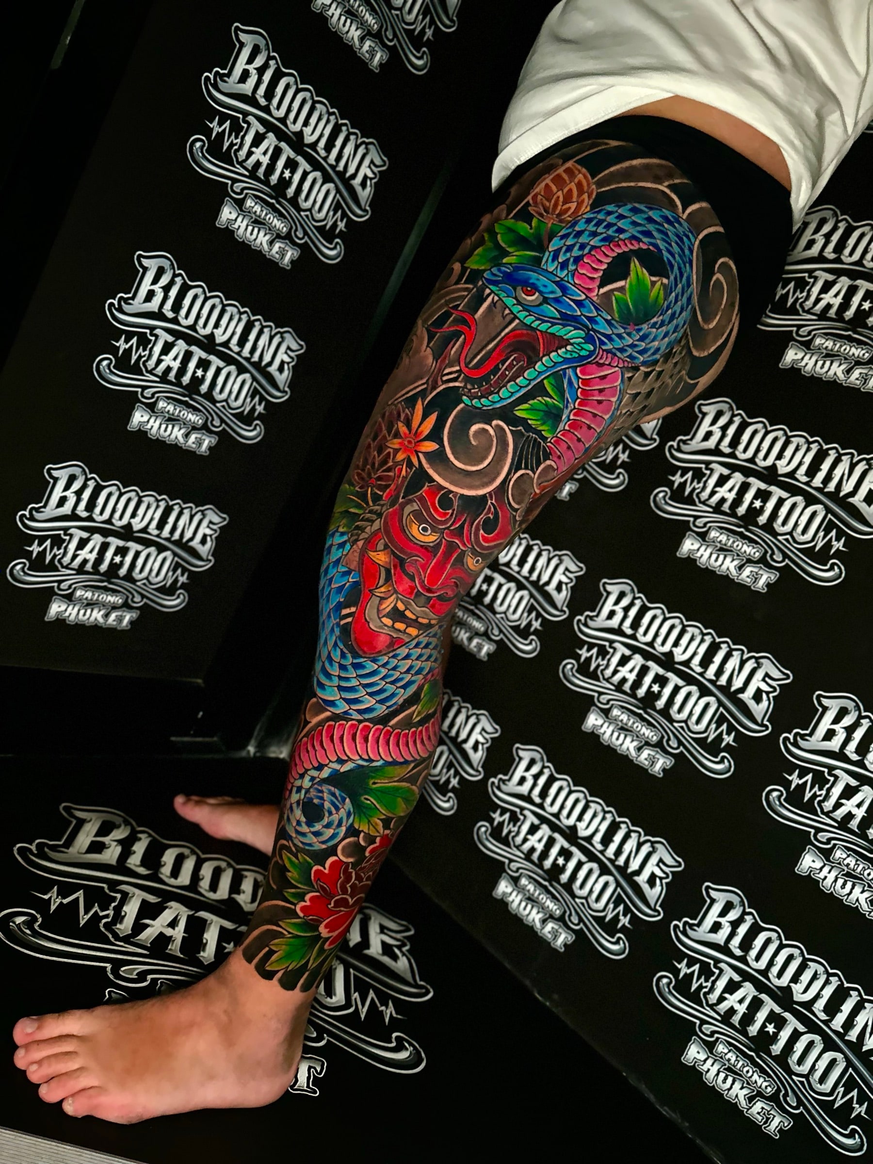 Japanese colour full-leg tattoo with a coiled blue and pink snake, a red Hannya mask, and floral accents in black wind-bar shading at Bloodline Tattoo Phuket.