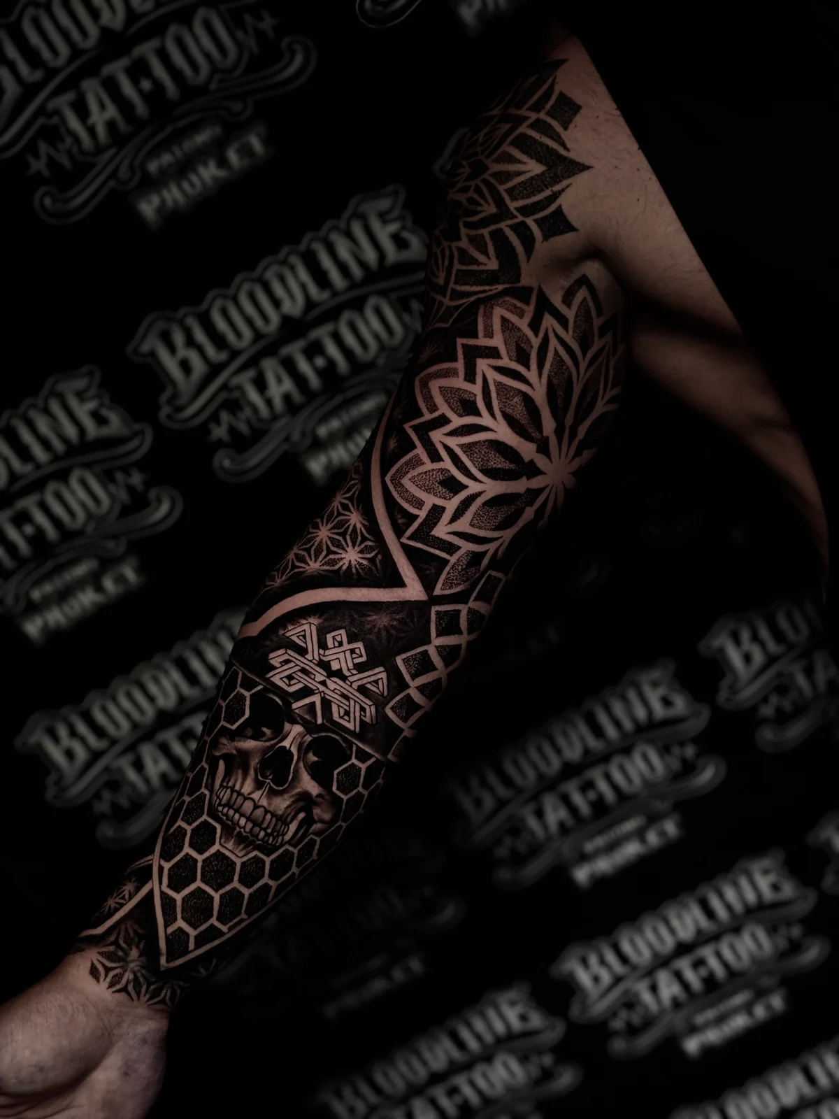 Mandala and geometric sleeve with shoulder lotus mandalas, a framed forearm skull, and honeycomb wrist patterns in black ink at Bloodline Tattoo Phuket.