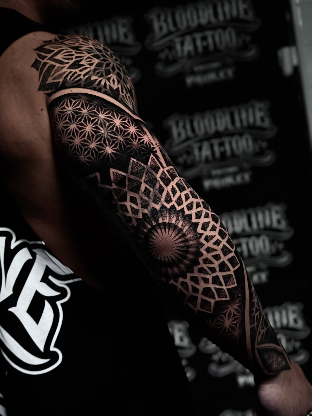 Black ink geometric mandala sleeve with a pointed shoulder mandala, Flower of Life upper arm fill, and a sunburst elbow focal point at Bloodline Tattoo Phuket.