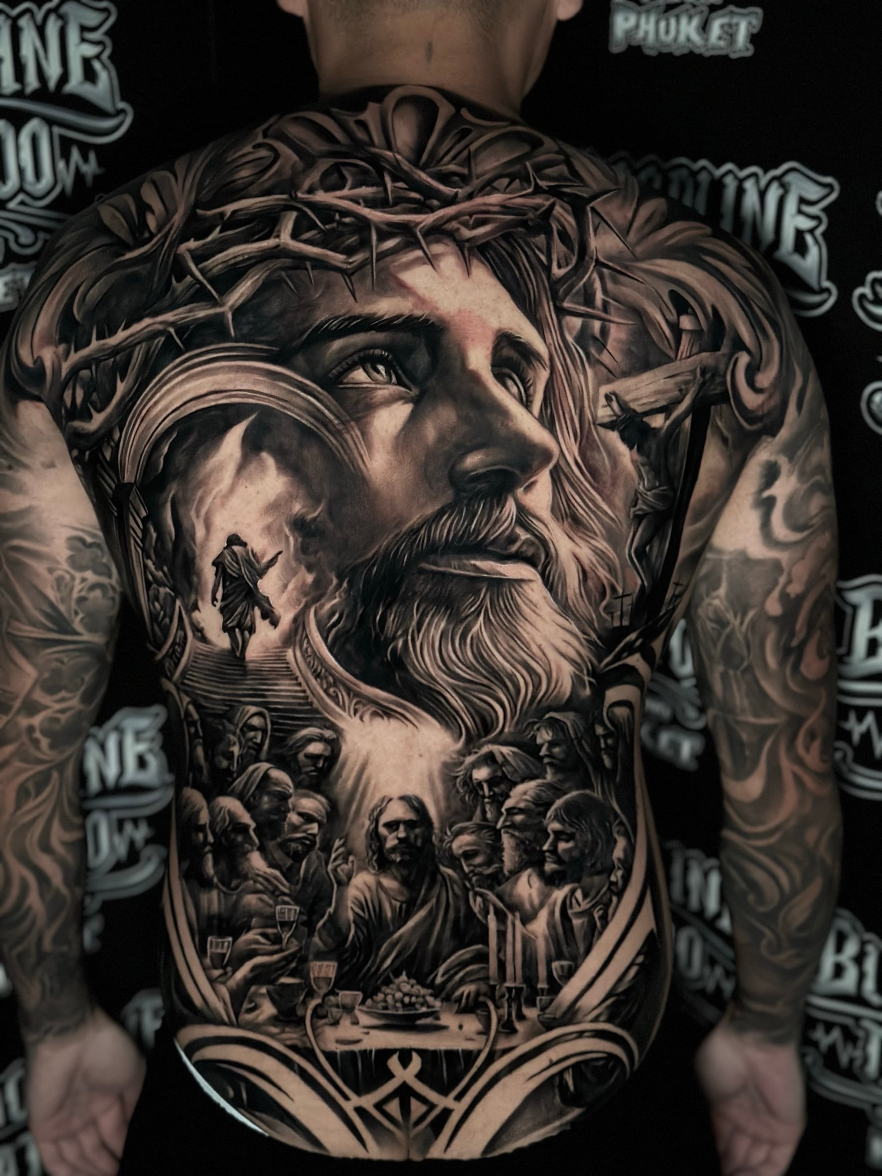 Large black and grey realism full back tattoo featuring Jesus with a crown of thorns, the Last Supper, the Crucifixion, and a figure ascending toward light at Bloodline Tattoo Phuket.
