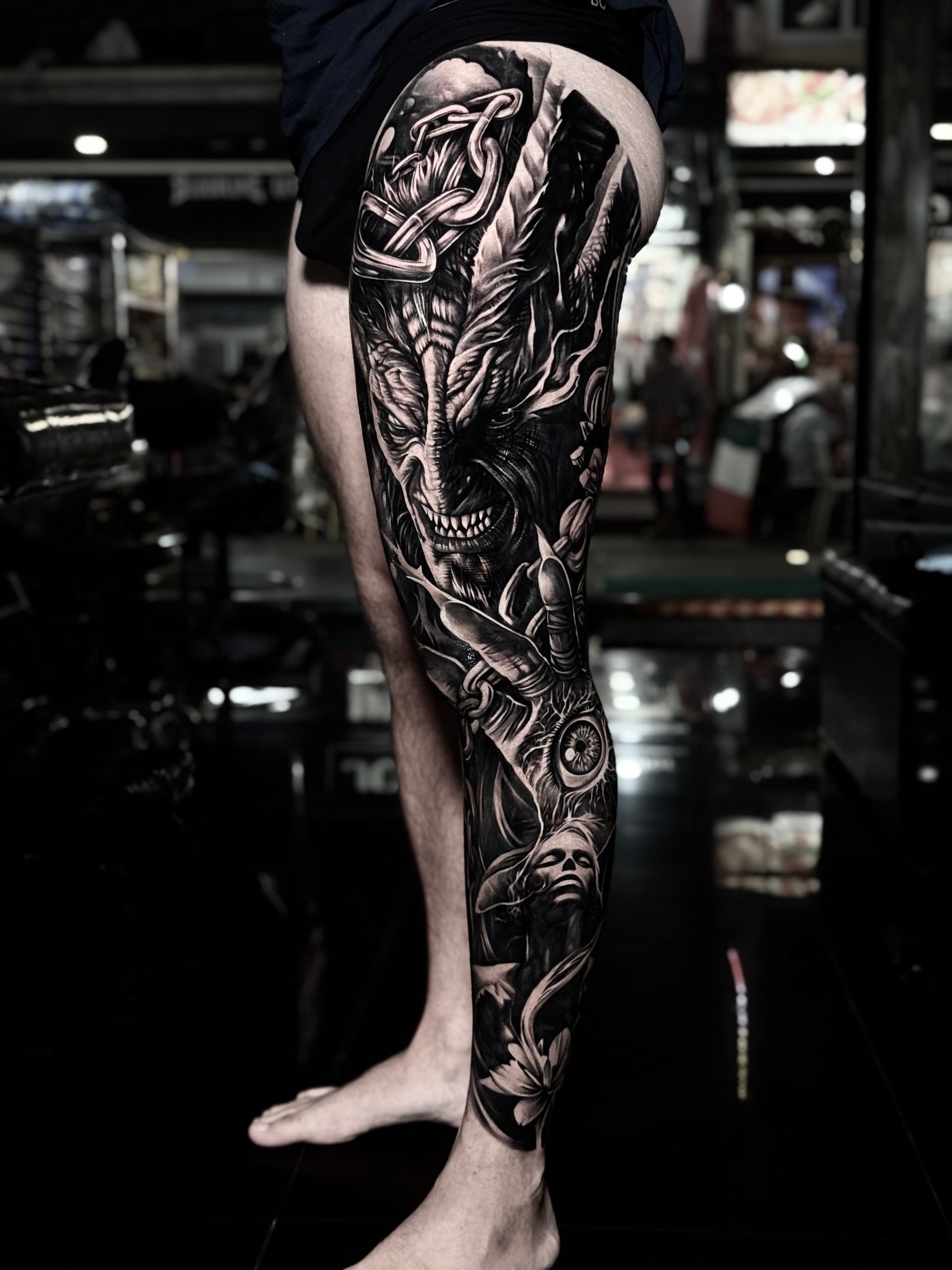Detailed black and grey realism full leg sleeve tattoo featuring a snarling demon, metal chains, a realistic eye in a clawed hand, a serene woman, and a lotus flower at Bloodline Tattoo Phuket.
