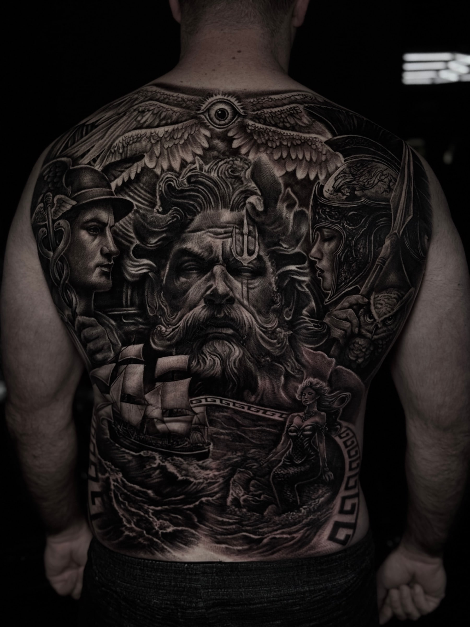 Highly detailed black and grey realism full back tattoo featuring Poseidon, winged all-seeing eye, Hermes, Athena with owl, sailing ship, mermaid, and Greek key border at Bloodline Tattoo Phuket.