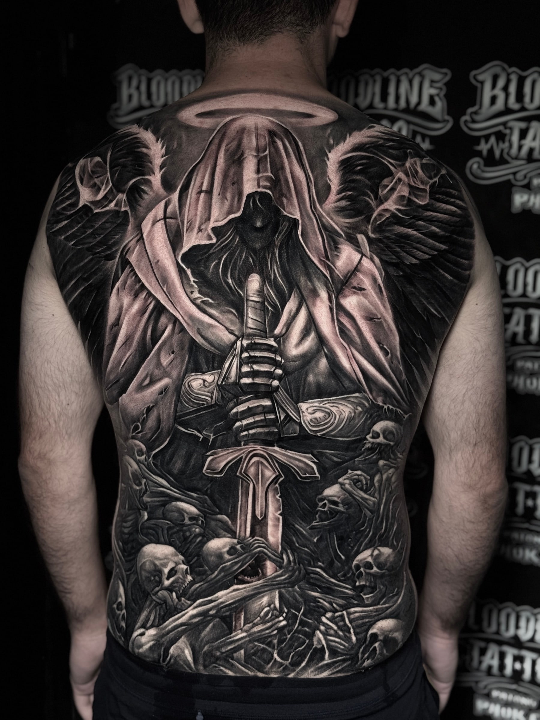 Detailed black and grey realism full back tattoo of a hooded winged angel holding a broadsword above a pile of skulls and reaching skeletal hands at Bloodline Tattoo Phuket.