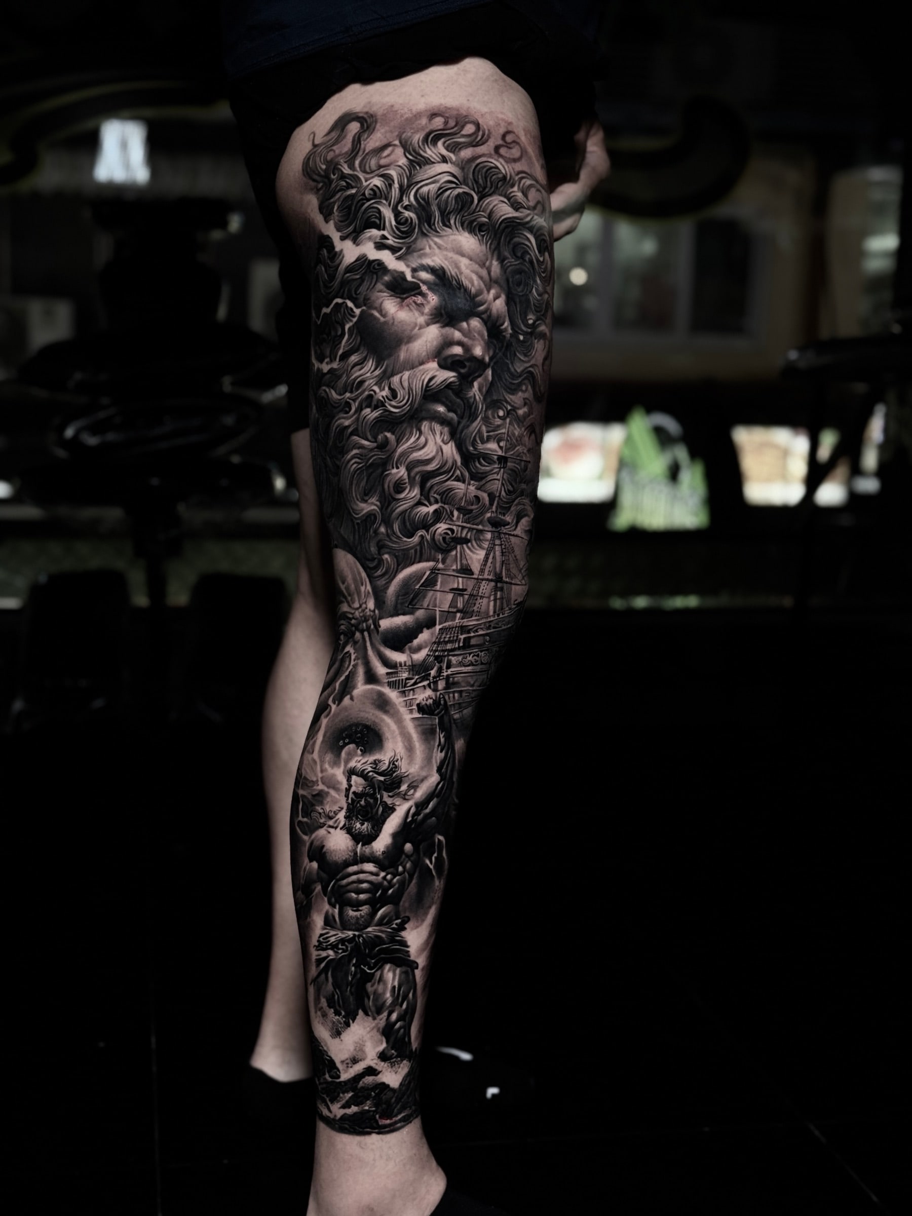 Detailed black and grey realism full leg sleeve tattoo featuring a Greek god portrait with lightning, a sailing galleon, and a muscular figure in stormy waves at Bloodline Tattoo Phuket.