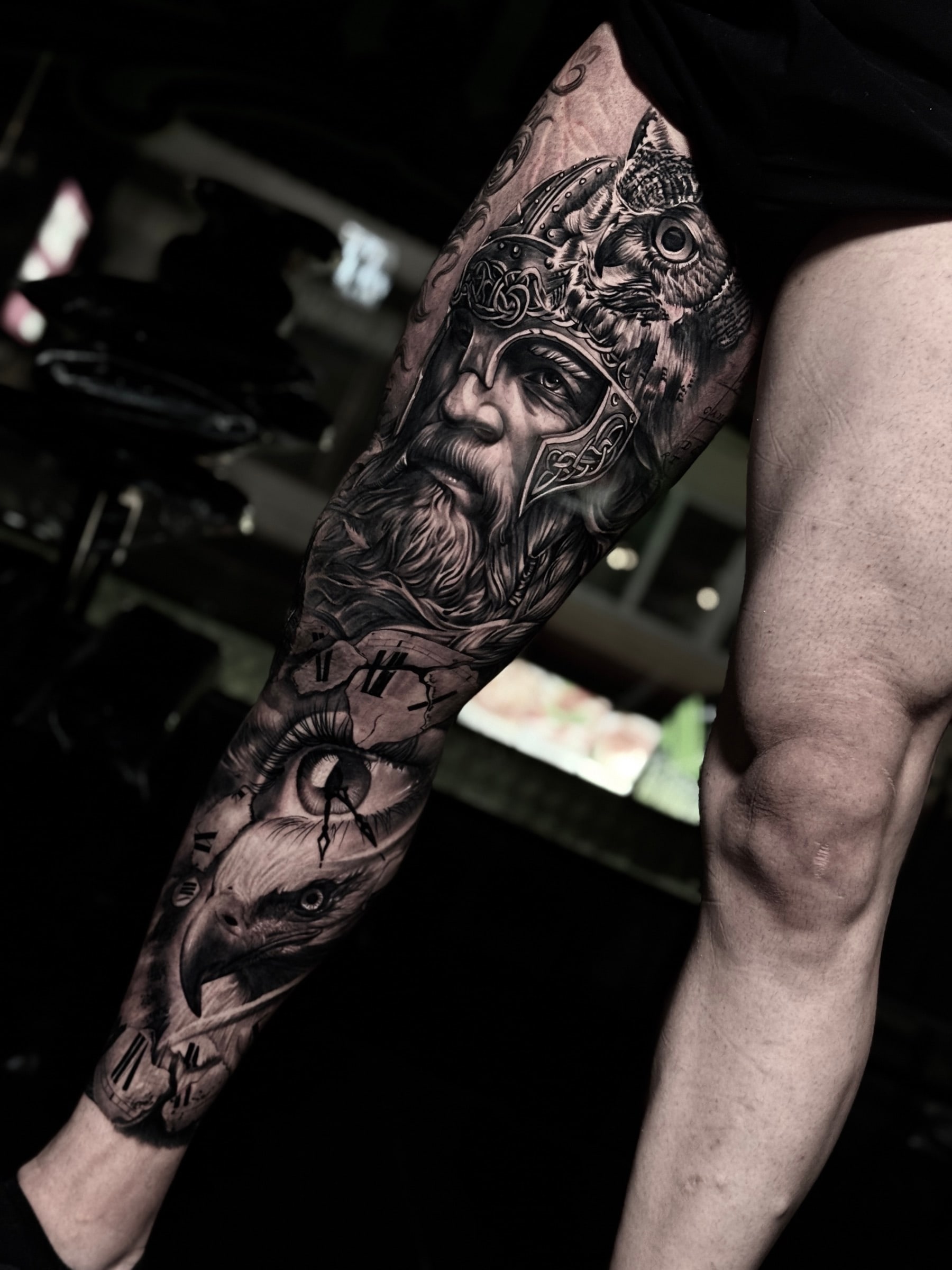 Detailed black and grey realism full leg sleeve tattoo featuring a Viking warrior in a knotwork helmet, an owl, a shattered clock with a realistic eye, and an eagle head at Bloodline Tattoo Phuket.