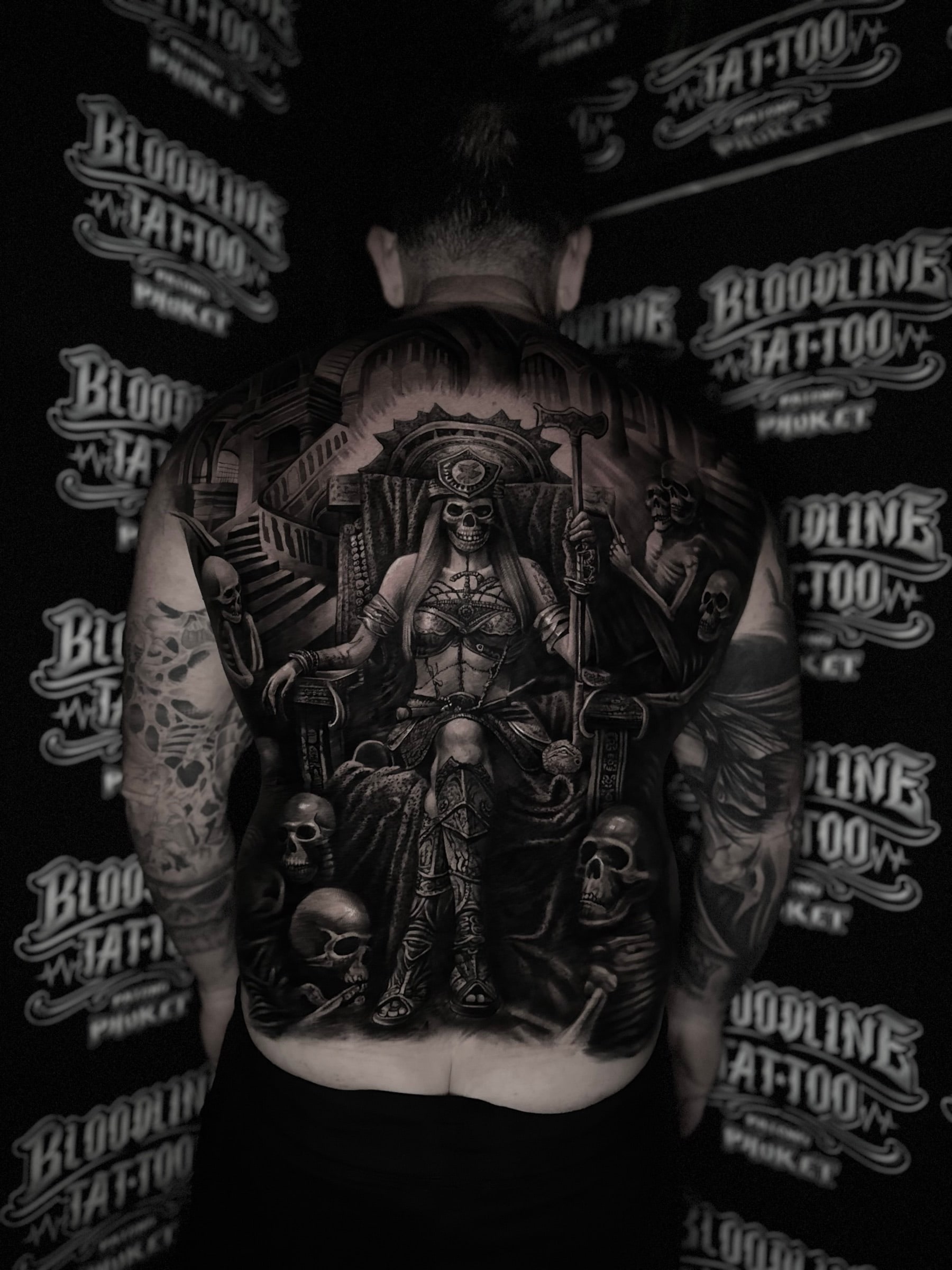 Detailed black and grey realism full back tattoo of a skull-faced warrior woman on a gothic throne with staff, skulls, and skeletal figure at Bloodline Tattoo Phuket.