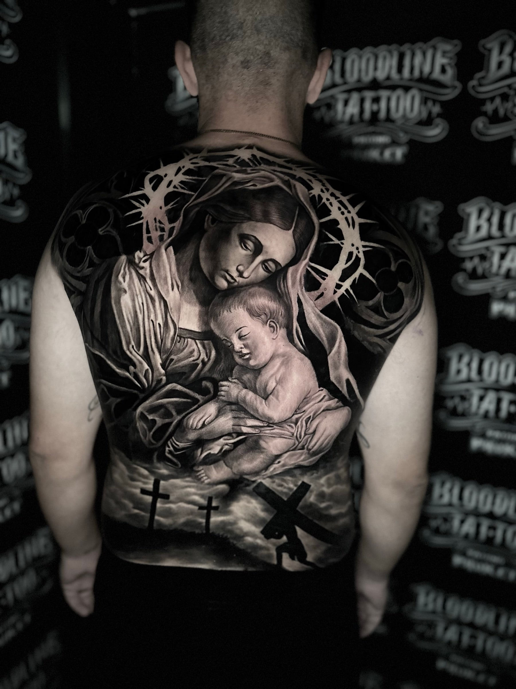 Detailed black and grey realism full back tattoo of the Virgin Mary holding the infant Jesus beneath a crown of thorns and Calvary crosses at Bloodline Tattoo Phuket.