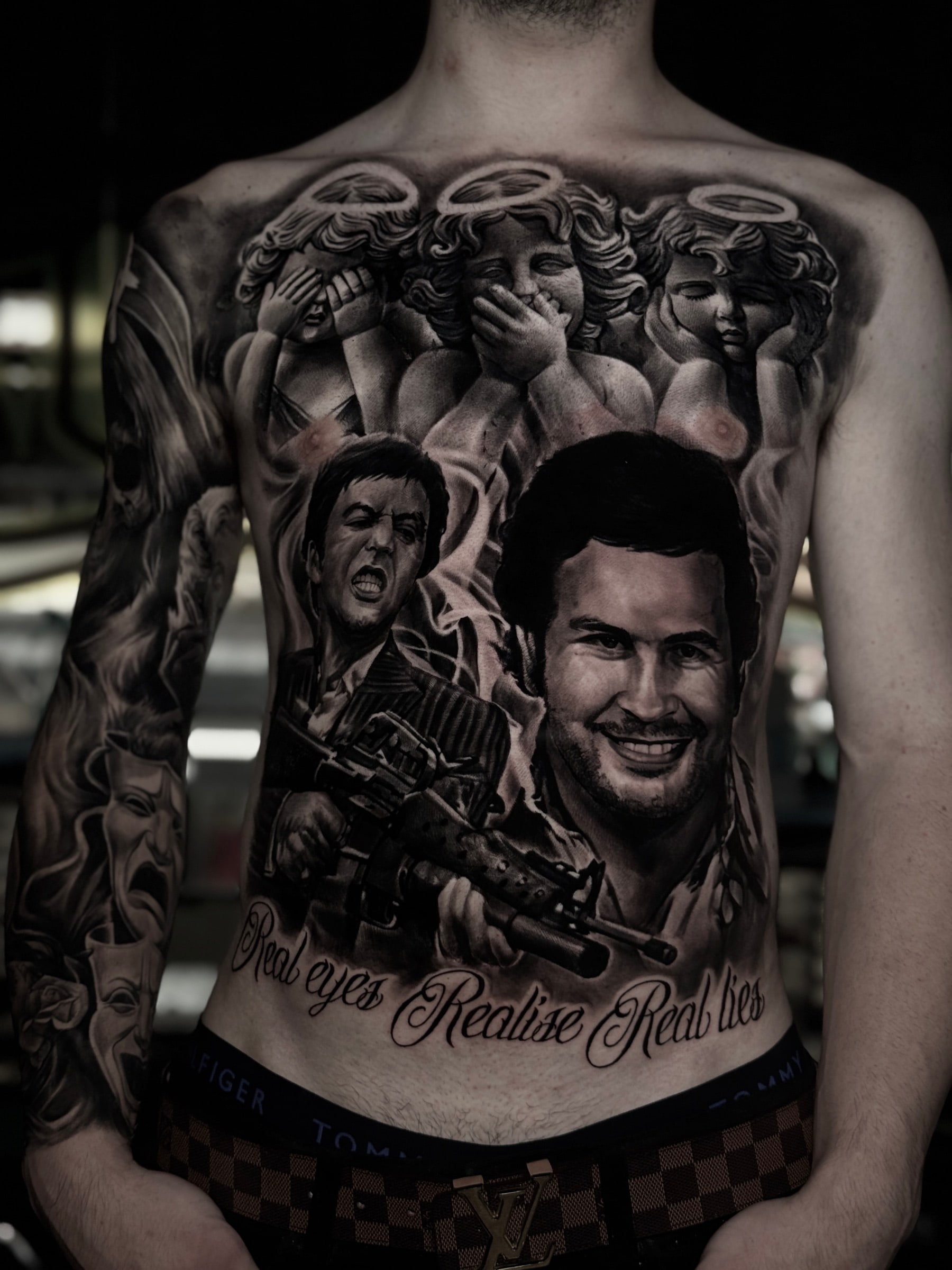 Detailed black and grey realism torso tattoo featuring cherubs, Tony Montana from Scarface, Pablo Escobar, and script reading Real eyes Realise Real lies at Bloodline Tattoo Phuket.