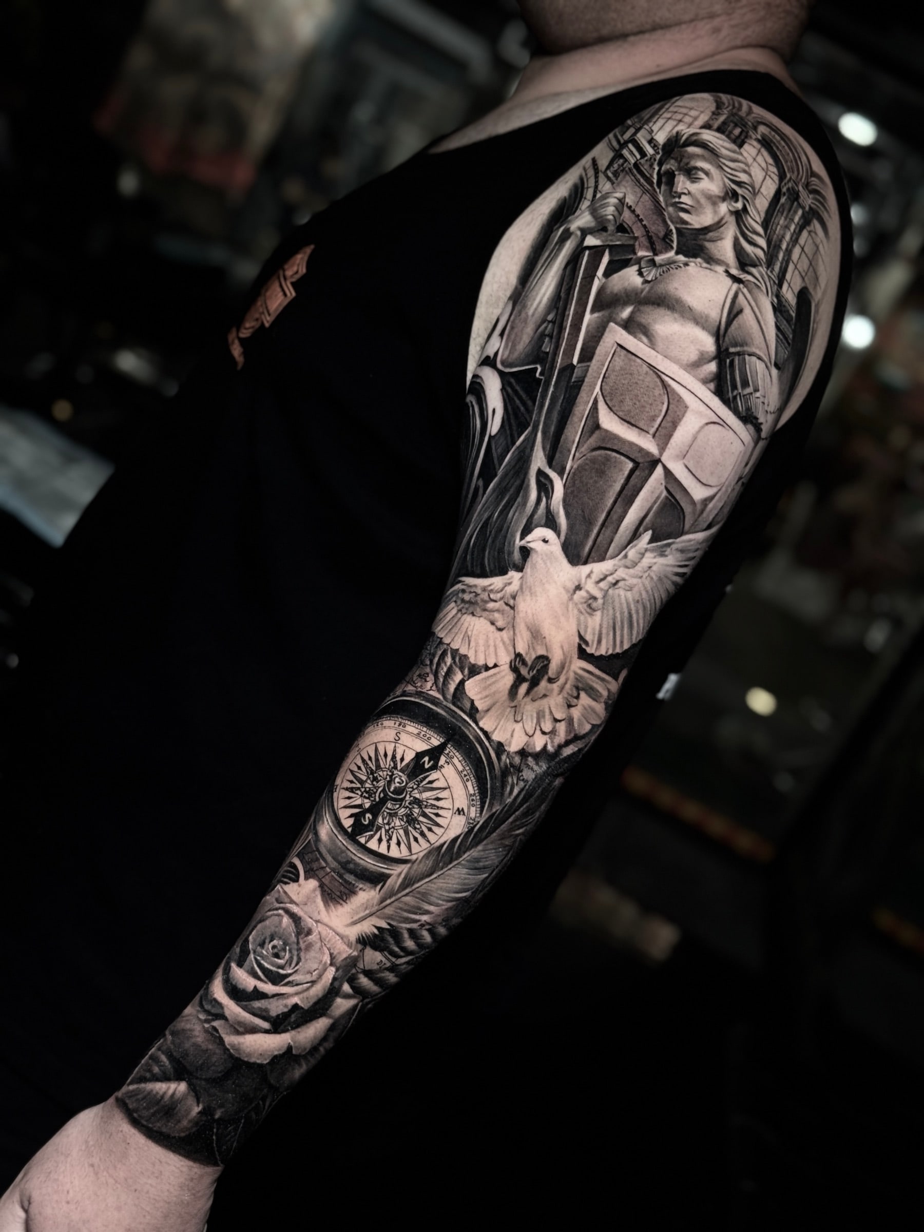 Detailed black and grey realism full sleeve tattoo featuring a classical warrior with cross shield, flying dove, compass rose, rose, and feather at Bloodline Tattoo Phuket.