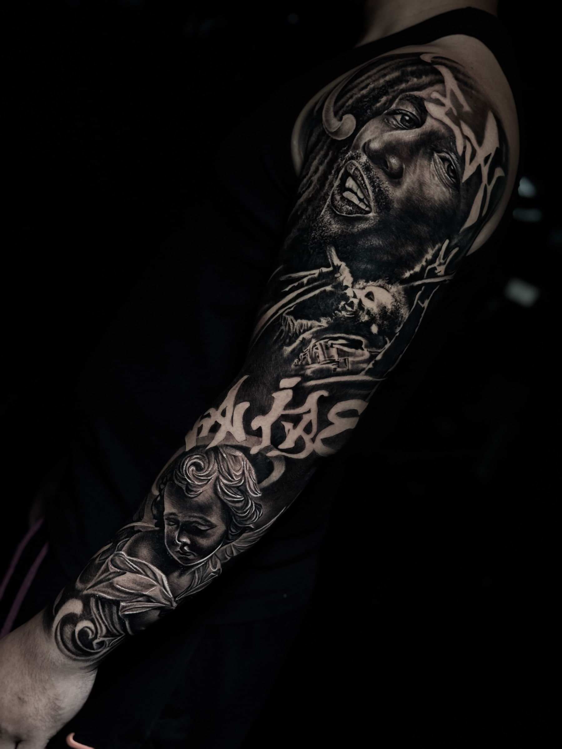 Detailed black and grey realism full sleeve tattoo featuring a Bob Marley portrait, skeletal scene, script lettering, and cherub angel at Bloodline Tattoo Phuket.